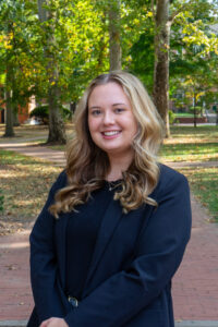 Anna is standing in front of The Class Gateway and smiling. She is wearing a black shirt and a black blazer.